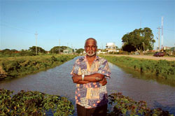 Vince Ramcharran stands near the river where he swam as a child growing up on Albion sugar cane plantation 30 miles from New Amsterdam, Guyana. To the right is the noisy cane factory. Credit: Leslie Latcha