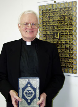 Fr. Dave Warren holding the Qur'an. Behind him is a poster featuring the 99 Names of God (Allah).